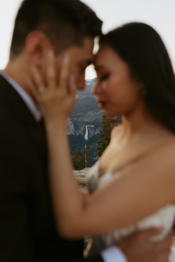 couples share a sweet moment on glacier point with a waterfall behind them as the focus of the photo 