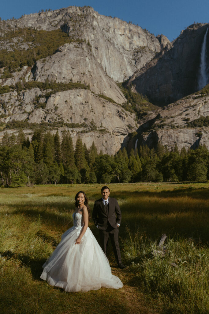 couple takes bride and grooms portraits in front of Yosemite falls in Cooks meadow 